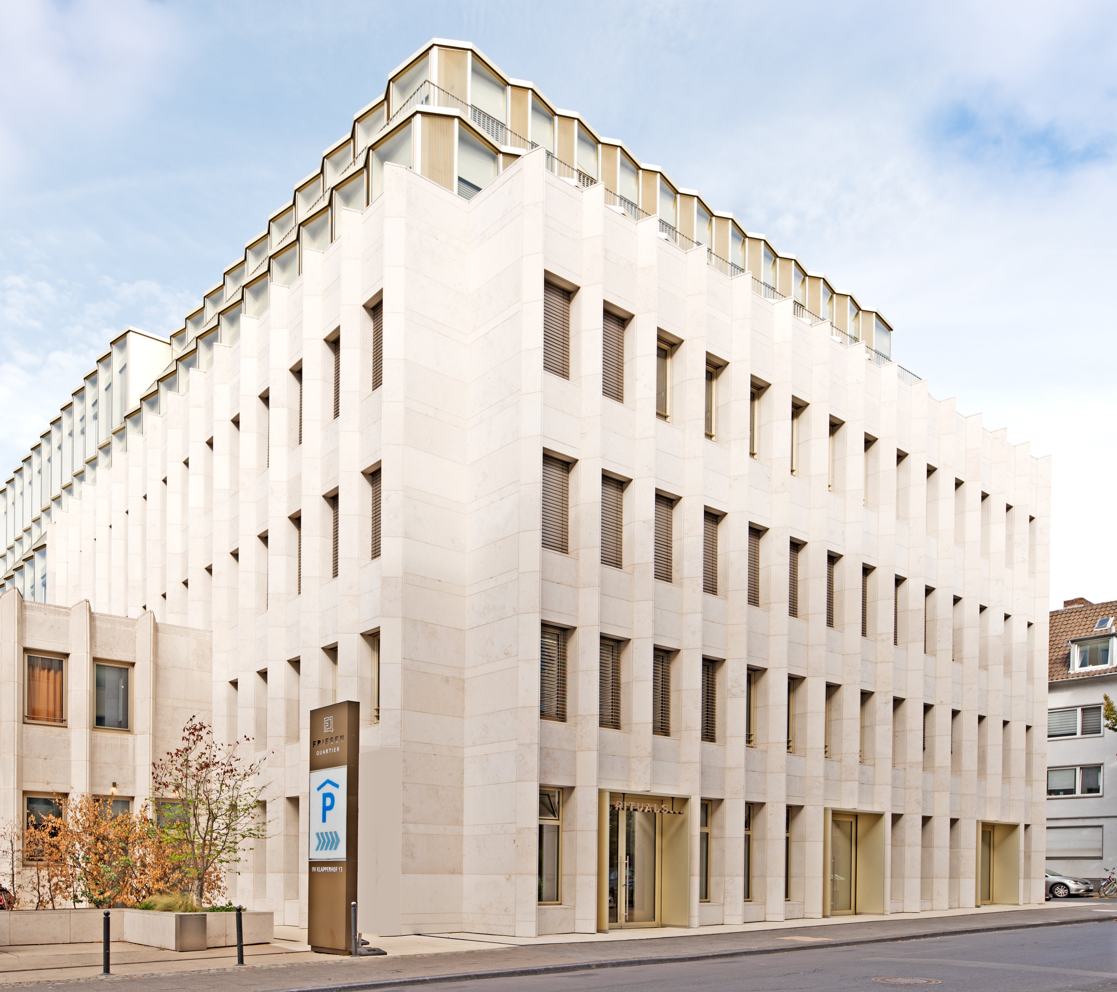 Saniertes Eckgebäude des Projekts Gerling Garden an der Kreuzung Im Klapperhof in Köln, das eine helle Steinfassade und markante, abgestufte Staffelgeschosse zeigt. Die moderne Architektur integriert sich harmonisch in die historische Umgebung.