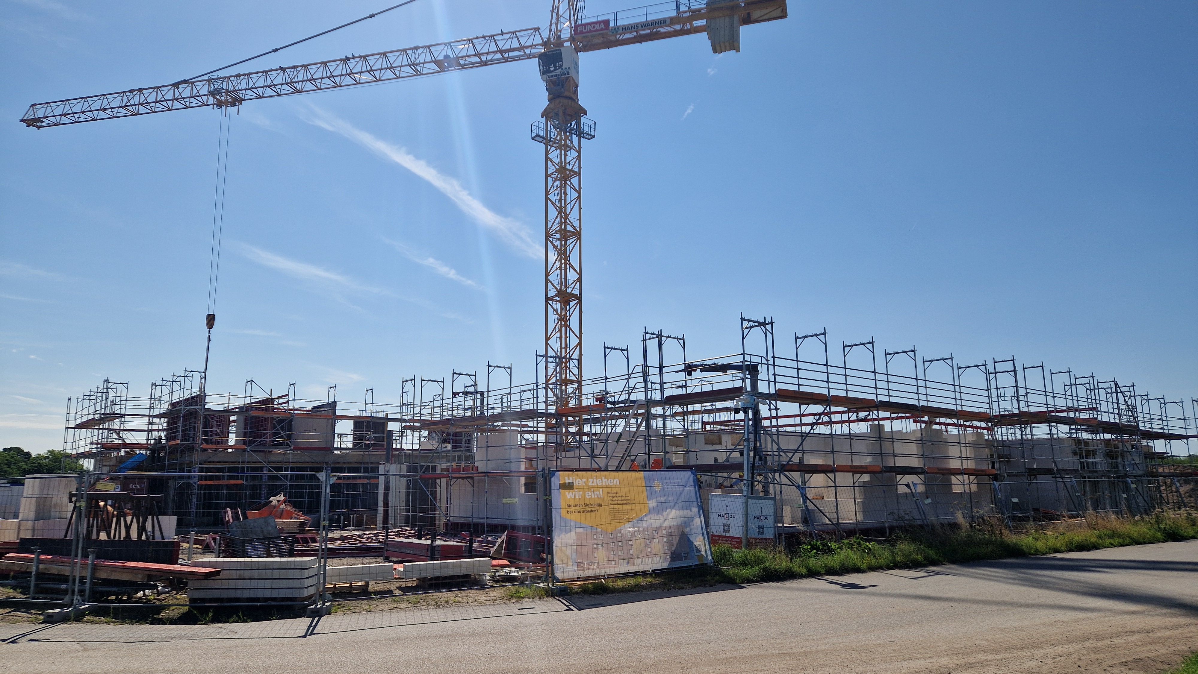 Rohbau eines mehrgeschossigen Hauses mit klar erkennbaren Betonelementen, großen Glasflächen und Gerüstteilen. Umgeben von Pflasterwegen, Rasen und frisch gepflanzten Sträuchern.

              