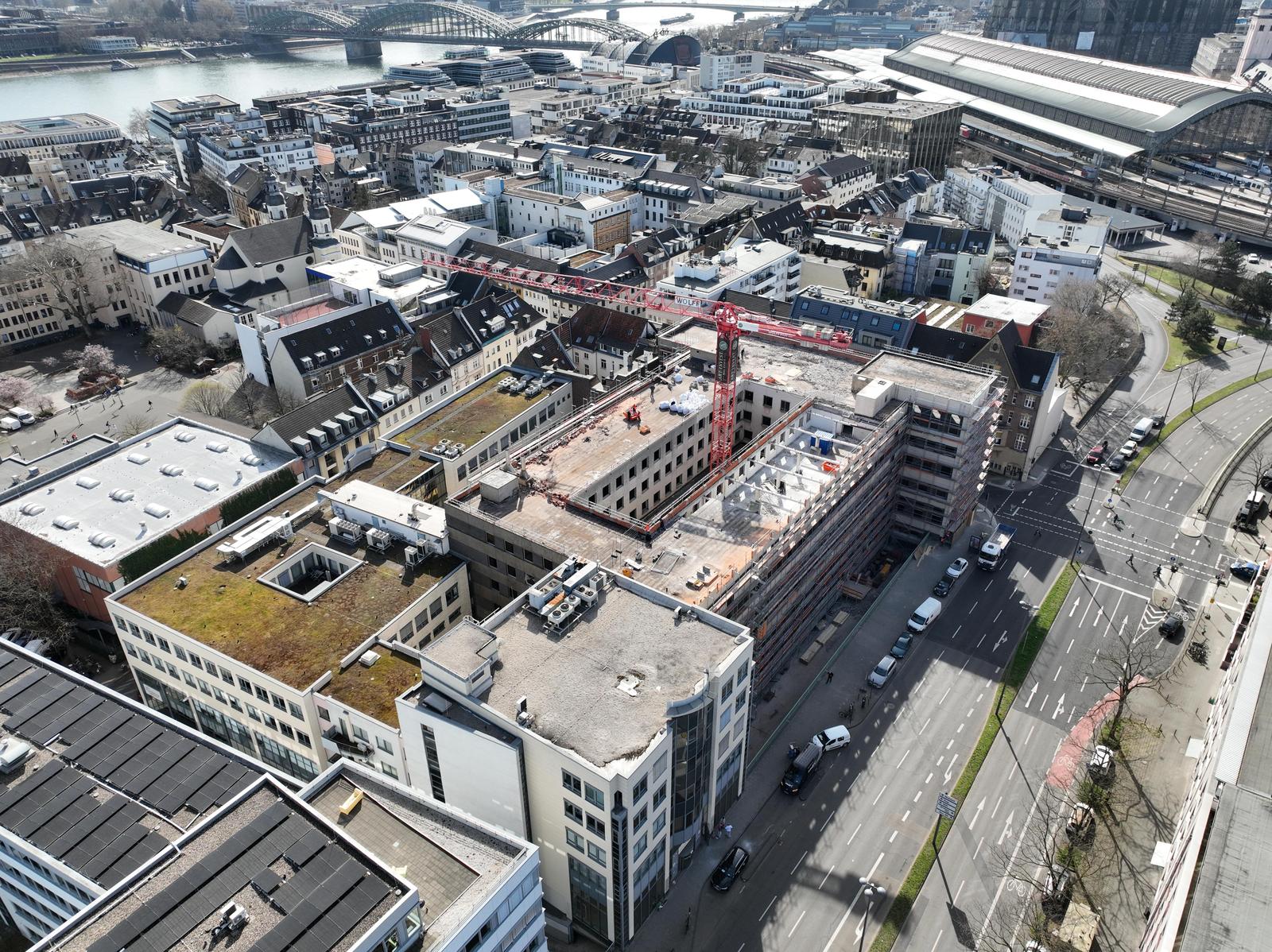 Visualisierung eines modernen Bürogebäudes an einer großen Kreuzung. Der Gebäudekomplex besteht aus einem weißen Eckgebäude mit Rundbögen und einem längeren Flügel mit vertikalen Fassadenelementen. Autos und Fußgänger auf der Straße, blauer Himmel mit Wolken.