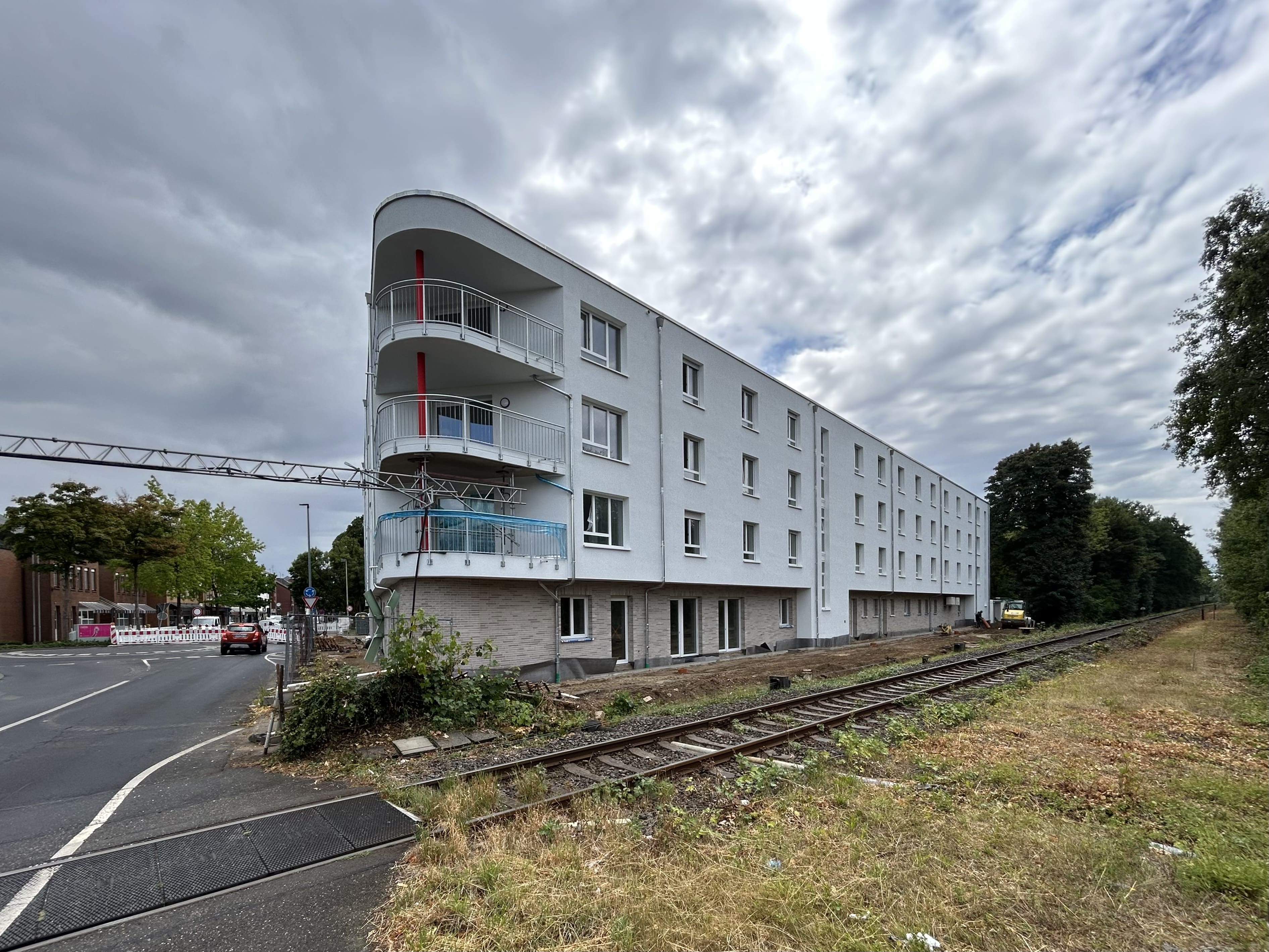 Luftbild einer Innenstadt mit Fluss und Brücken im Hintergrund. Im Zentrum eine Baustelle mit rotem Kran, umgeben von mehrgeschossigen Gebäuden. Das Bauprojekt liegt an einer Hauptstraße, zahlreiche Autos und Lieferwagen parken am Straßenrand.