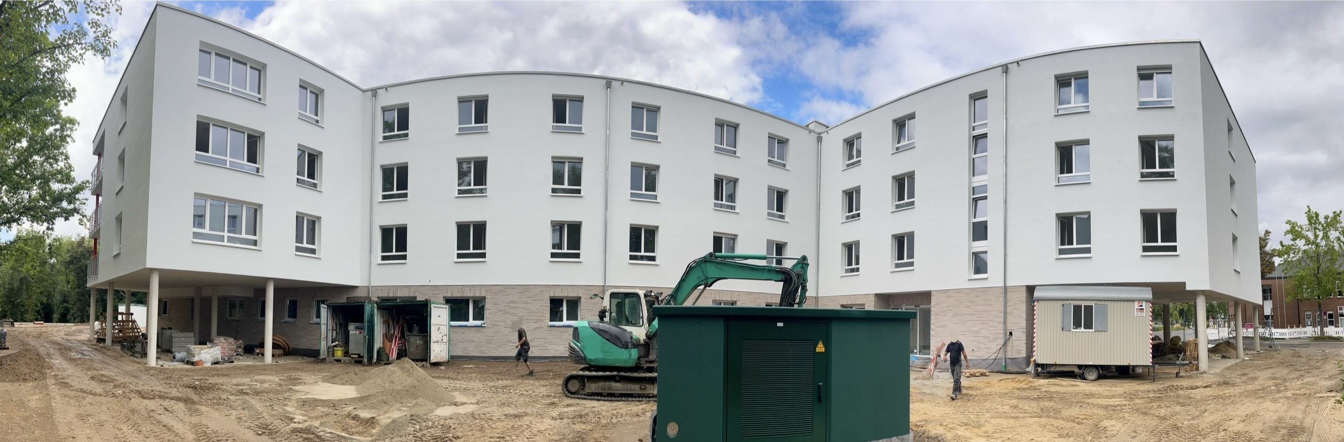 Luftbild einer Innenstadt mit Fluss und Brücken im Hintergrund. Im Zentrum eine Baustelle mit rotem Kran, umgeben von mehrgeschossigen Gebäuden. Das Bauprojekt liegt an einer Hauptstraße, zahlreiche Autos und Lieferwagen parken am Straßenrand.