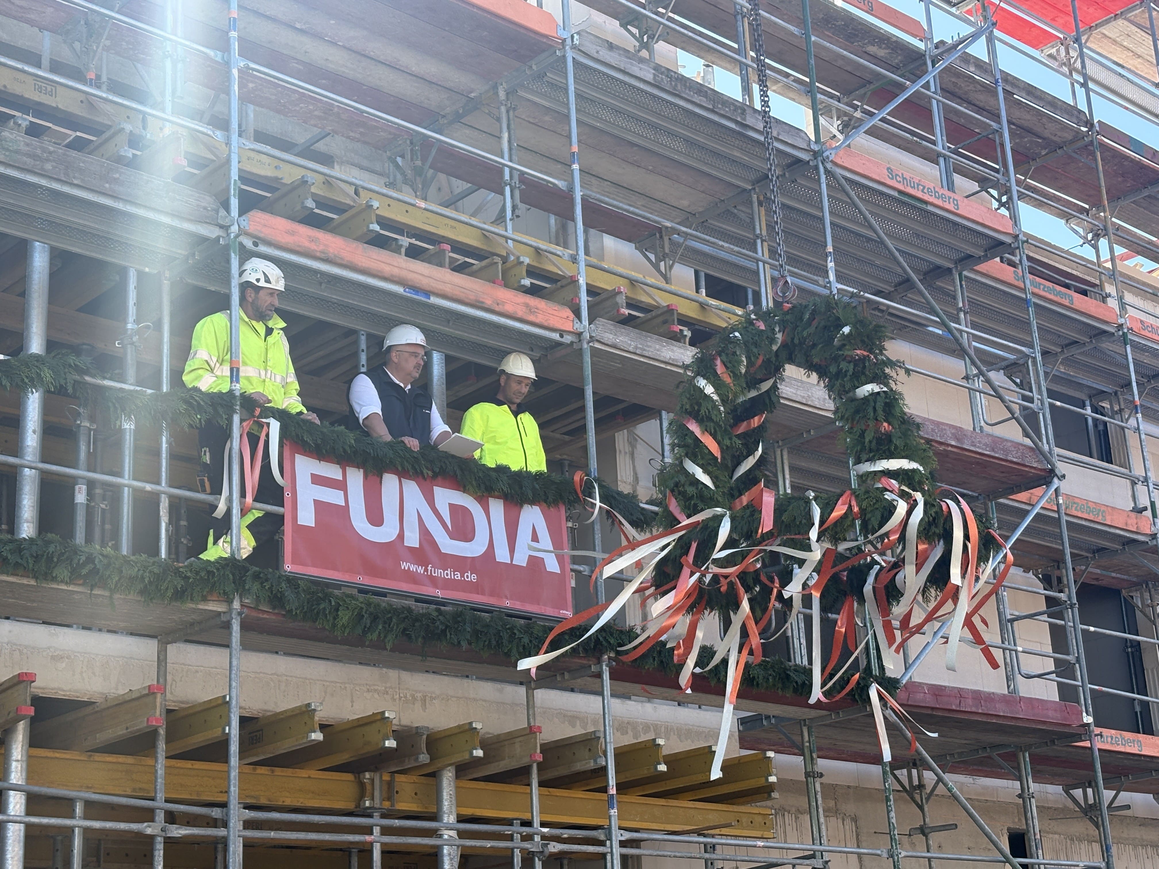 Drei FUNDIA-Bauarbeiter mit Helmen stehen stolz auf dem Gerüst hinter einem roten FUNDIA-Banner, daneben hängt der geschmückte Richtkranz.