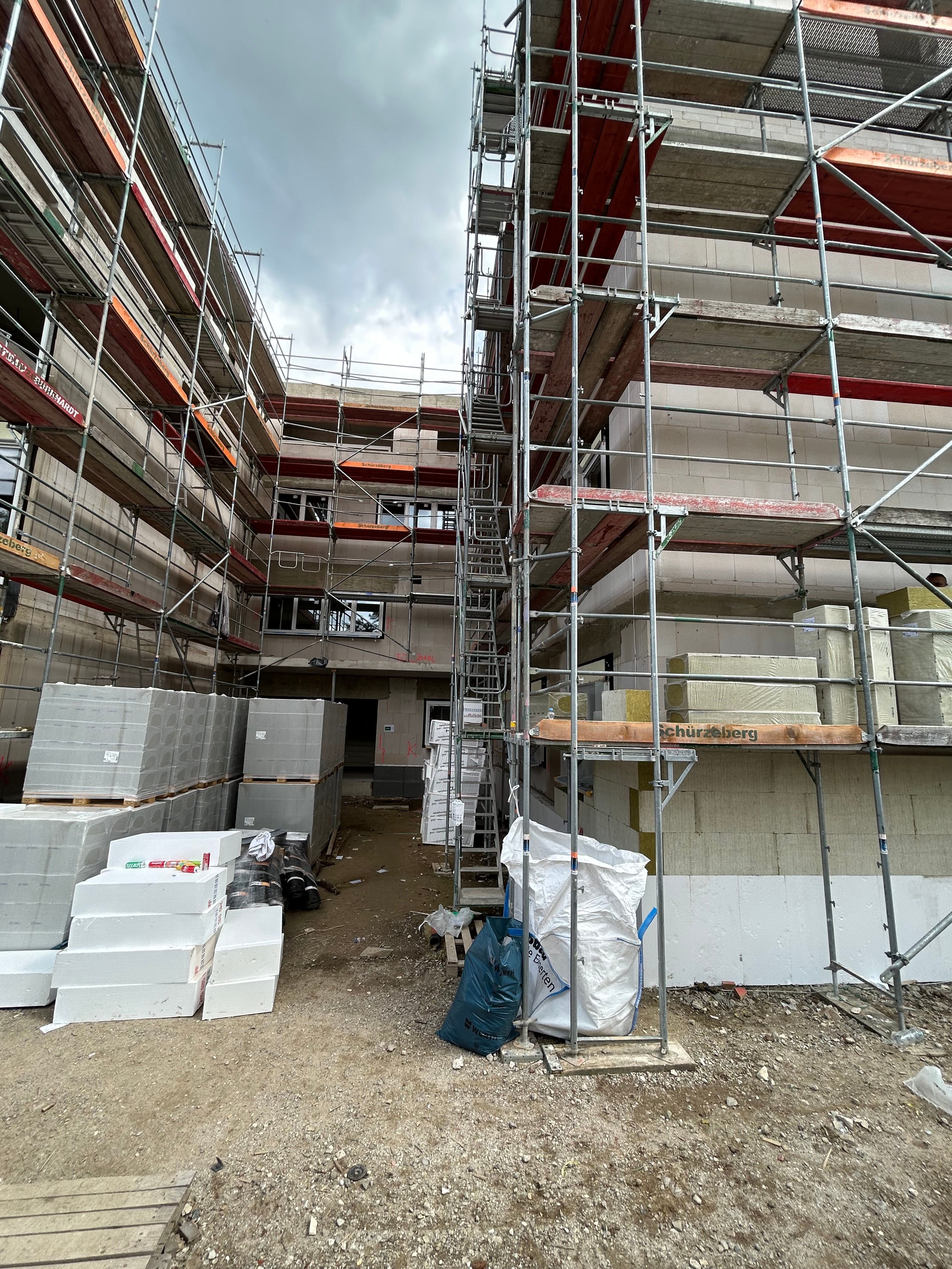 Baustelle eines modernen Wohngebäudes mit sichtbaren Betondecken, Gerüsten und offenen Fensteröffnungen. Im Vordergrund verlaufen Gehwege, Rasenflächen und junge Bäume entlang der Baustelle.