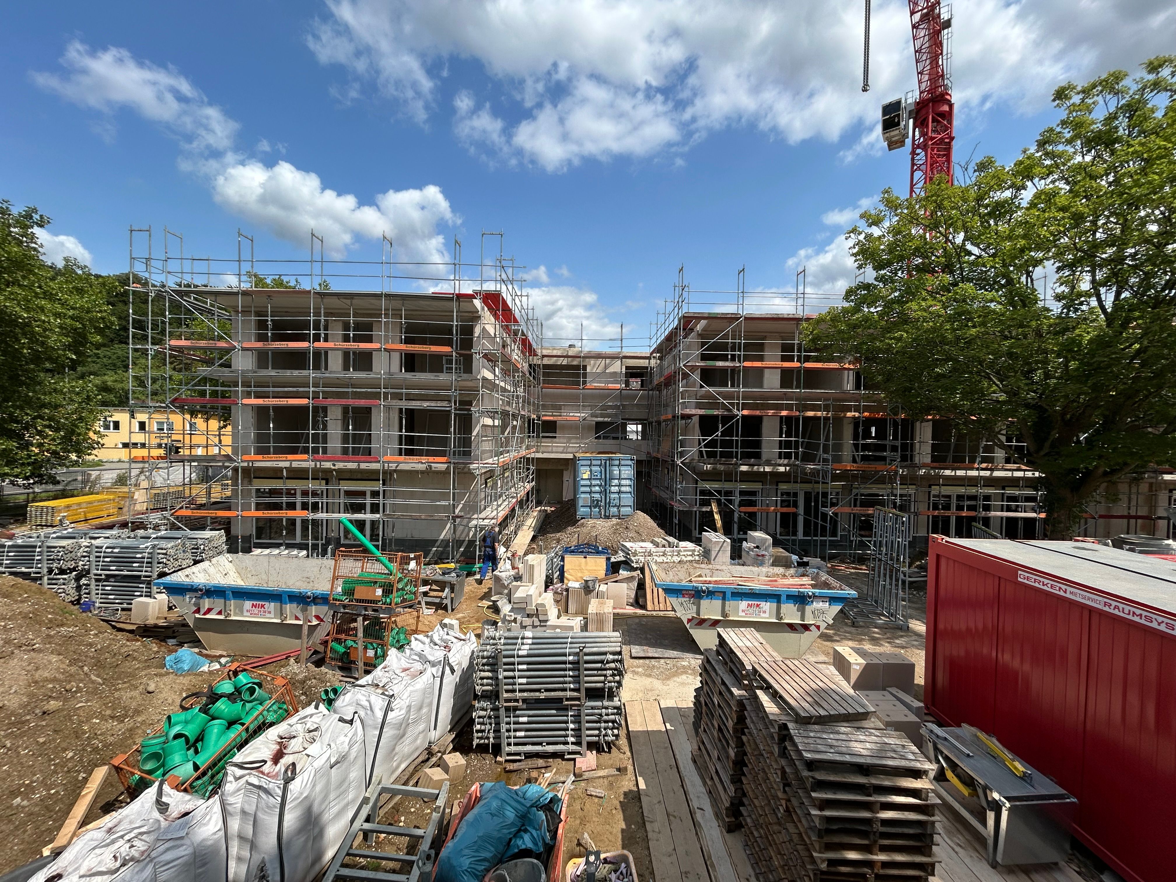 Baustelle eines modernen Wohngebäudes mit sichtbaren Betondecken, Gerüsten und offenen Fensteröffnungen. Im Vordergrund verlaufen Gehwege, Rasenflächen und junge Bäume entlang der Baustelle.