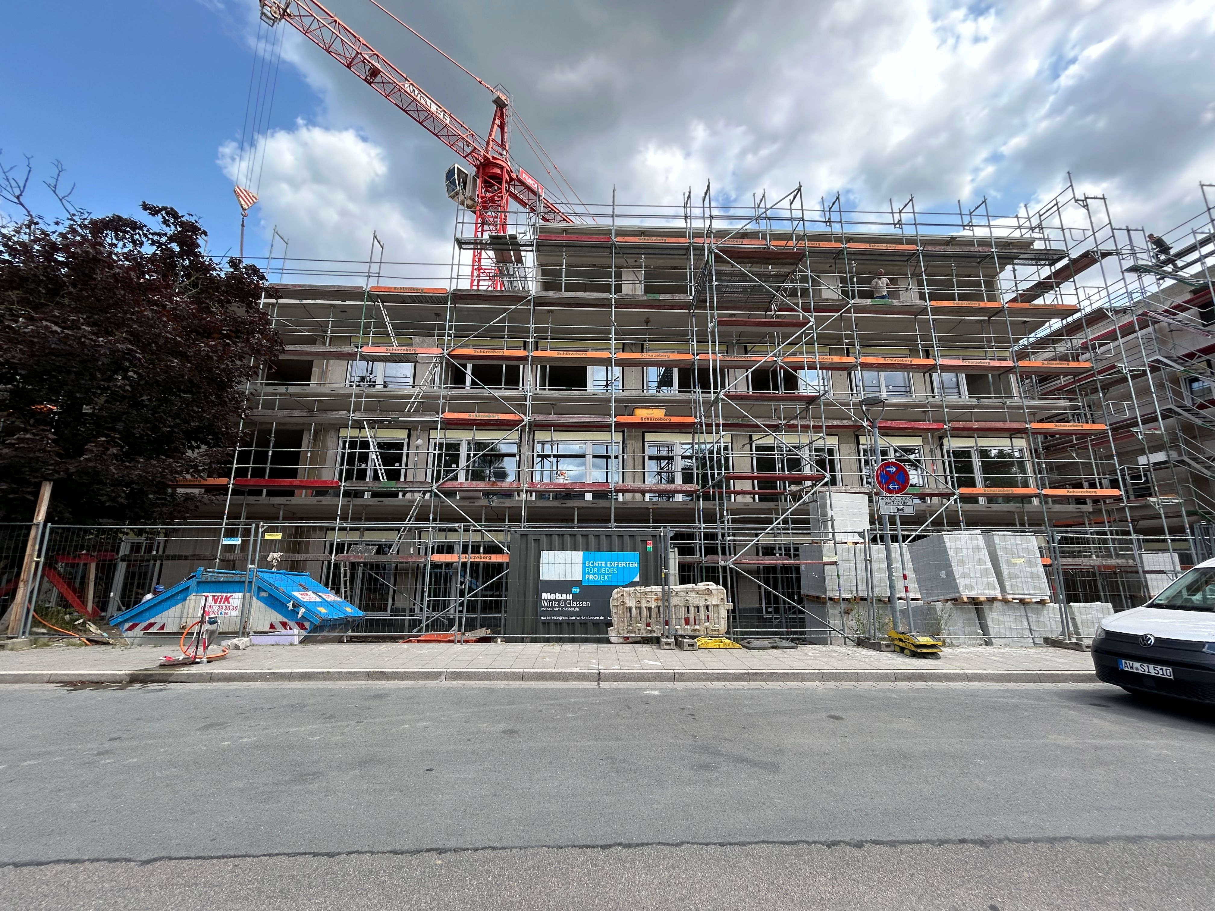 Rohbau eines mehrgeschossigen Hauses mit klar erkennbaren Betonelementen, großen Glasflächen und Gerüstteilen. Umgeben von Pflasterwegen, Rasen und frisch gepflanzten Sträuchern.

              