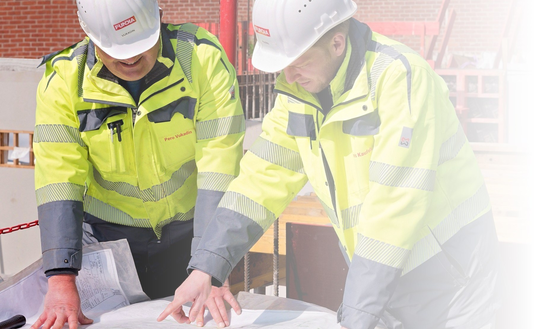 Zwei Fundia Mitarbeiter mit Arbeitskleidung und Helmen sprechen über ausgebreiteten Plänen